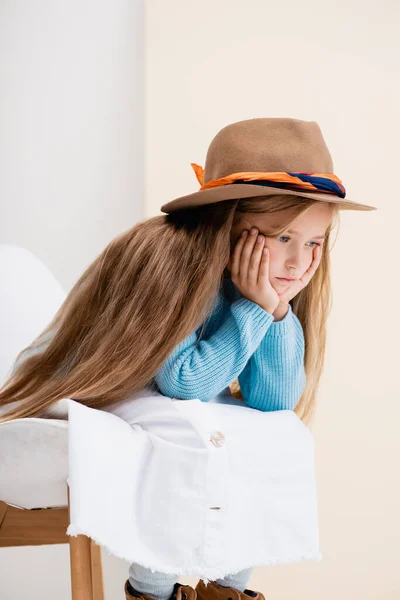 Triste fille blonde à la mode en chapeau marron, jupe blanche et pull bleu assis sur une chaise près du mur beige — Photo de stock