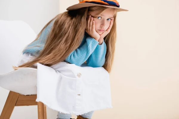 Sad fashionable blonde girl in brown hat, white skirt and blue sweater sitting on chair near beige wall — Stock Photo