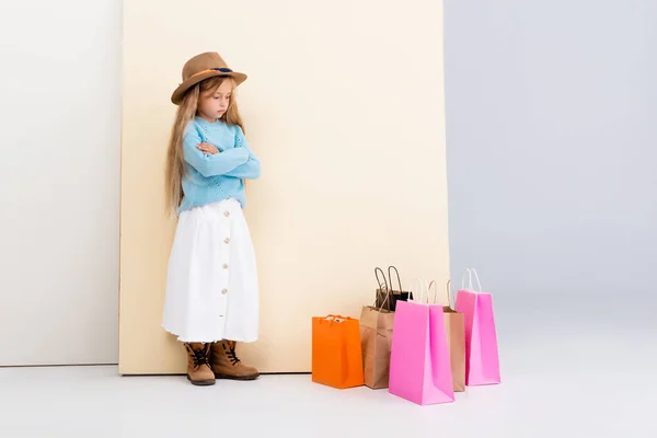 Triste chica rubia de moda en sombrero marrón y botas, falda blanca y suéter azul cerca de coloridas bolsas de compras y la pared beige - foto de stock