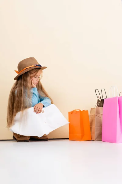 Triste chica rubia de moda en sombrero marrón y botas, falda blanca y suéter azul sentado cerca de coloridas bolsas de compras y la pared beige - foto de stock