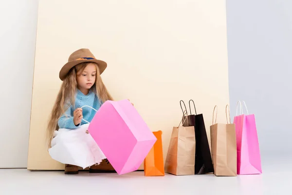 Fashionable blonde girl in brown hat and boots, white skirt and blue sweater looking inside colorful shopping bag near beige wall — Stock Photo