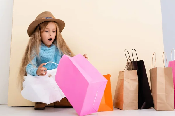 Schockiertes modisches blondes Mädchen mit braunem Hut und Stiefeln, weißem Rock und blauem Pullover schaut in bunte Einkaufstasche nahe beiger Wand — Stockfoto