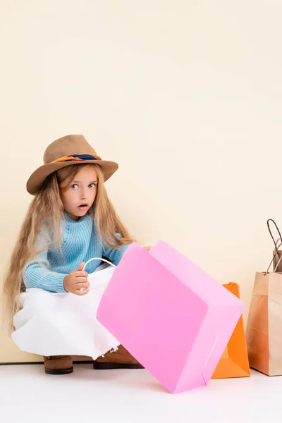 Shocked fashionable blonde girl in brown hat and boots, white skirt and blue sweater looking inside colorful shopping bag near beige wall — Stock Photo