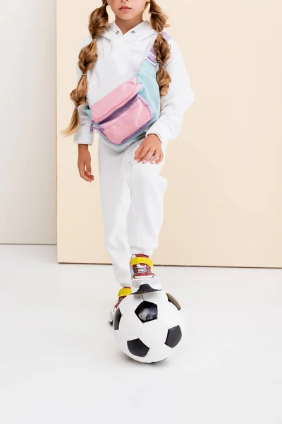 Обрезанный вид на блондинку в спортивной форме позируя с футбольным мячом на бежевом и белом фоне — стоковое фото