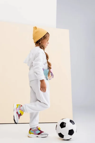 Side view of blonde girl in sportswear posing with soccer ball on beige and white background — Stock Photo