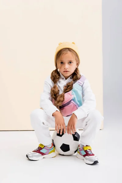 Menina loira em sportswear posando com bola de futebol no fundo bege e branco — Fotografia de Stock