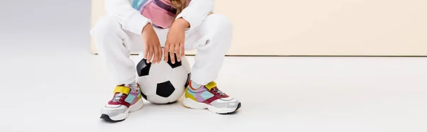 Recortado vista de chica en ropa deportiva posando con pelota de fútbol sobre fondo beige y blanco, tiro panorámico - foto de stock