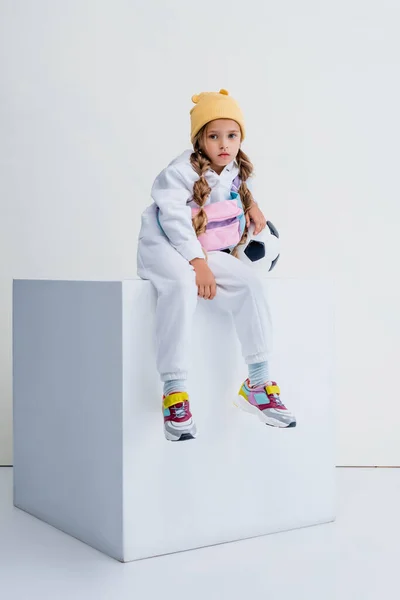Rubia chica en ropa deportiva sentado en cubo con pelota de fútbol aislado en blanco - foto de stock