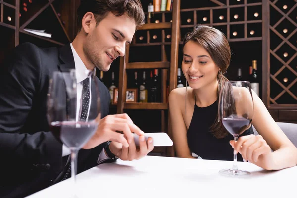 Enfoque selectivo del hombre en traje mostrando smartphone a novia con copa de vino en restaurante - foto de stock