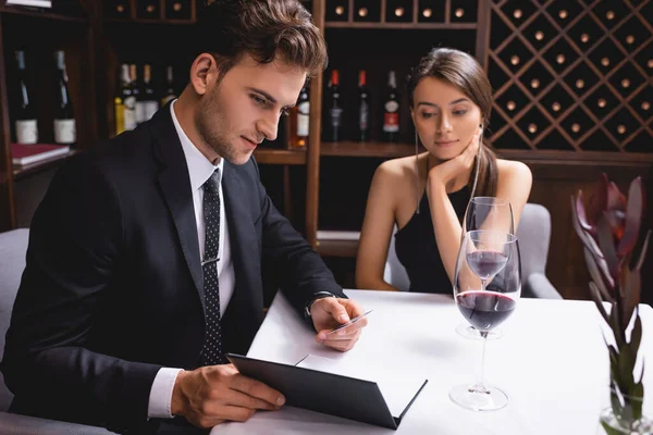Selective focus of man in suit looking at menu near glasses of wine during dating with girlfriend in restaurant — Stock Photo