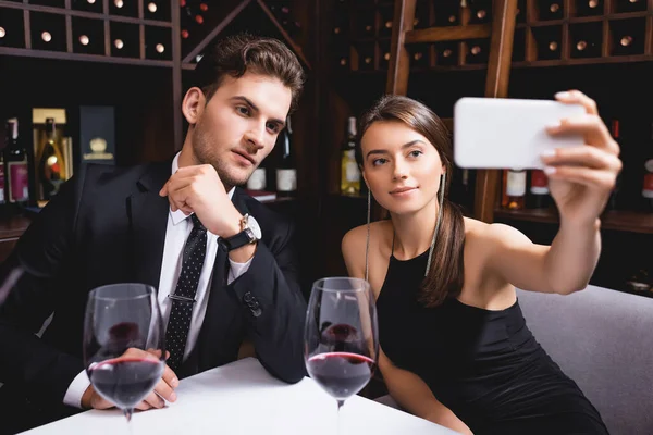 Foco seletivo de mulher elegante tomando selfie perto de namorado e copos de vinho no restaurante — Fotografia de Stock