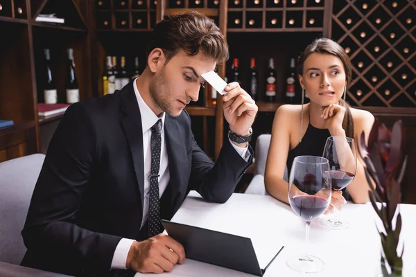 Concentration sélective de l'homme coûteux en costume tenant la carte de crédit tout en regardant le menu près des verres de vin et petite amie dans le restaurant — Photo de stock