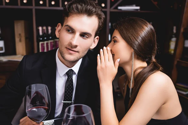 Selective focus of woman whispering to boyfriend in suit near glasses of wine in restaurant — Stock Photo