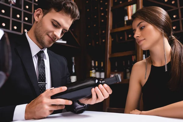 Выборочный фокус мужчины в костюме, держащего бутылку вина возле элегантной подружки в ресторане — стоковое фото