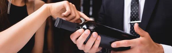 Encabezado del sitio web de la mujer que apunta a la botella de vino cerca de novio en traje en el restaurante - foto de stock