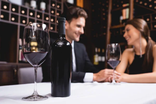 Enfoque selectivo de botella y copa de vino en la mesa cerca de pareja joven cogidos de la mano durante citas en el restaurante - foto de stock