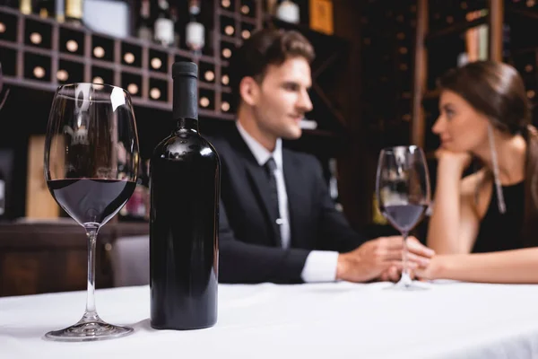 Enfoque selectivo de botella y copa de vino en la mesa cerca de pareja joven cogidos de la mano en el restaurante - foto de stock