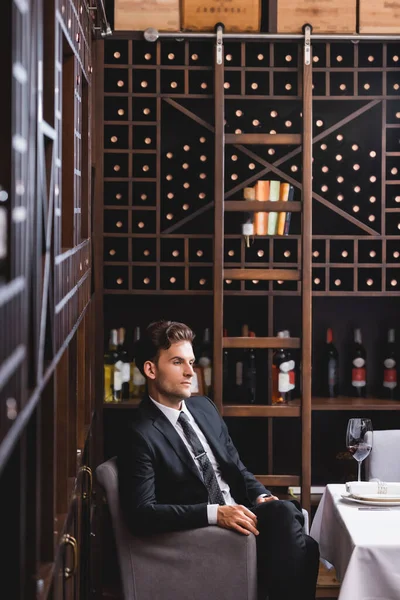 Selektiver Fokus eines jungen Mannes im Anzug, der in der Nähe von Weinglas und Regalen mit Weinflaschen im Restaurant sitzt — Stockfoto