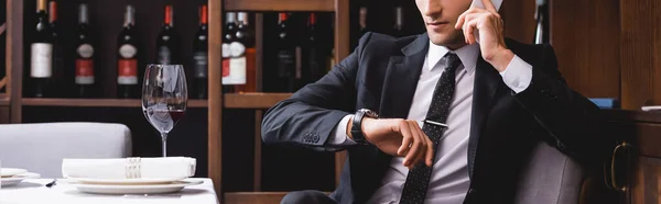 Panoramic shot of man in suit talking on smartphone and checking time on wristwatch near glass of wine in restaurant — Stock Photo