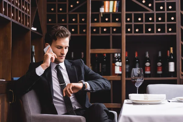 Selektiver Fokus des Mannes im Anzug, der mit dem Smartphone spricht und im Restaurant bei einem Glas Wein auf die Armbanduhr blickt — Stockfoto