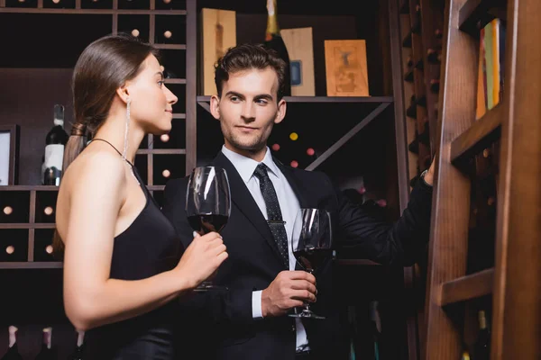 Casal elegante segurando copos de vinho enquanto olha para prateleiras com garrafas no restaurante — Fotografia de Stock