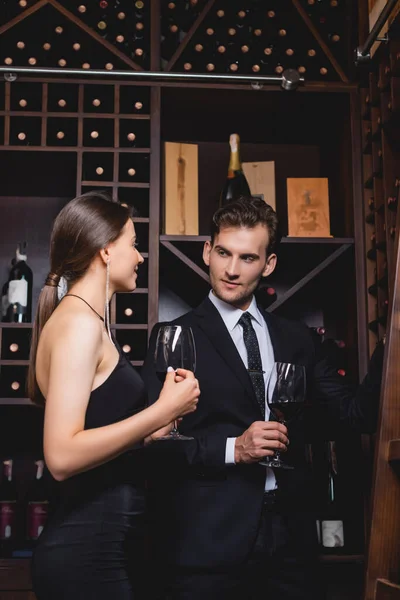 Hombre de traje sosteniendo copa de vino cerca de mujer elegante y bastidores con botellas en el restaurante - foto de stock