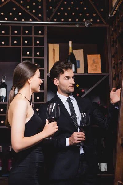 Mann im Anzug zeigt mit der Hand auf Regal mit Flaschen neben eleganter Frau mit Glas Wein im Restaurant — Stockfoto