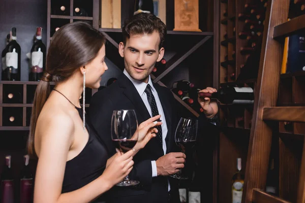 Selective focus of man in formal wear taking bottle from rack near elegant girlfriend with glass of wine — Stock Photo