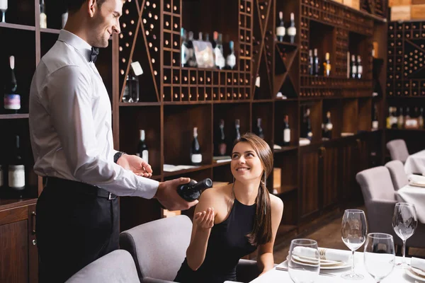 Selective focus of young woman pointing with hand near sommelier with bottle of wine in restaurant — Stock Photo