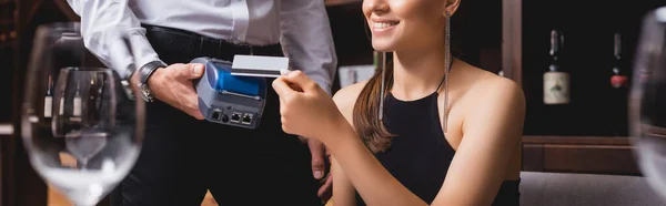 Panoramic shot of young woman paying with credit card to waiter with payment terminal in restaurant — Stock Photo