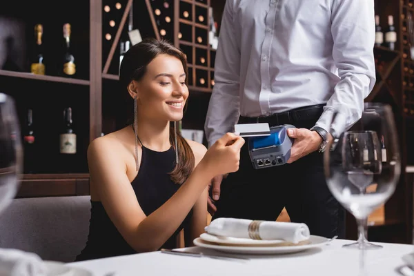 Selektiver Fokus der eleganten Frau mit Kreditkarte in der Nähe des Kellners mit Zahlungsterminal im Restaurant — Stockfoto