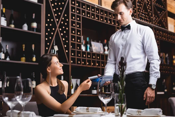Selective focus of young woman holding credit card near waiter in formal wear with payment terminal in restaurant — Stock Photo