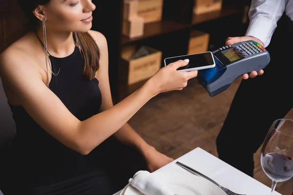 Vue recadrée de femme élégante payant avec smartphone pour serveur avec terminal de paiement dans le restaurant — Photo de stock