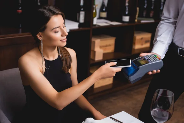 Concentration sélective de la jeune femme élégante payant avec téléphone portable près du serveur avec terminal de paiement dans le restaurant — Photo de stock