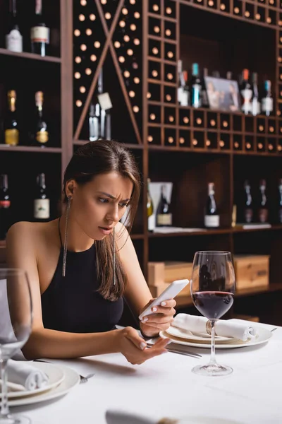 Selective focus of confused woman using smartphone while sitting near glass of wine in restaurant — Stock Photo