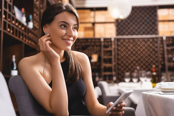 Selektiver Fokus einer eleganten Frau, die wegschaut, während sie ihr Smartphone im Restaurant hält — Stockfoto