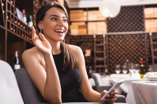 Enfoque selectivo de la mujer elegante agitando la mano mientras sostiene el teléfono inteligente en el restaurante - foto de stock