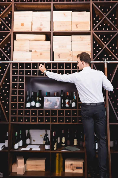 Visão traseira do sommelier em pé na escada enquanto empacotando garrafa de vinho do rack — Fotografia de Stock