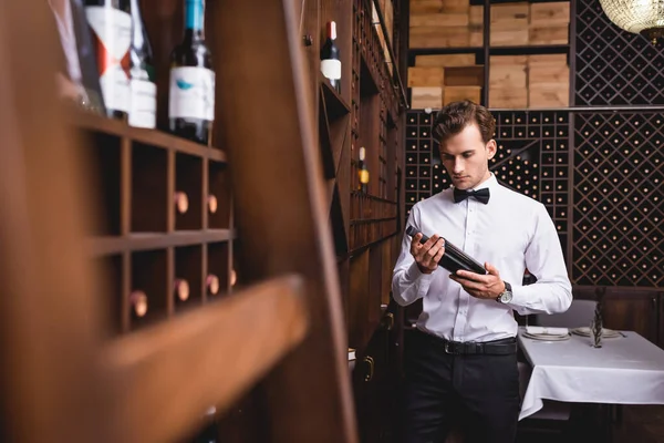 Selektiver Fokus des Sommeliers in formeller Kleidung, der eine Flasche Wein in der Nähe von Regalen im Restaurant hält — Stockfoto