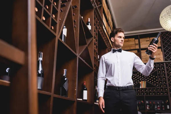 Выборочный фокус молодого сомелье в рубашке и галстуке-бабочке с бутылкой вина в ресторане — стоковое фото