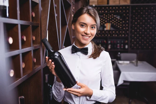 Selective focus of sommelier holding bottle of wine and looking at camera near racks — Stock Photo