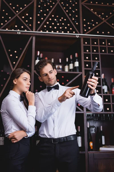 Sommelier señalando con la hormiga del dedo botella de vino cerca de colega pensativo en el restaurante - foto de stock