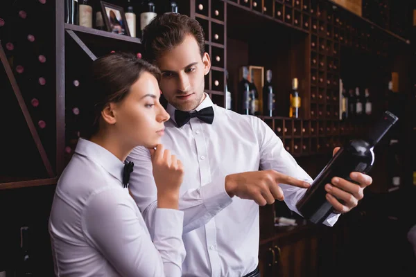 Selective focus of sommelier pointing at bottle of wine near thoughtful colleague in restaurant — Stock Photo