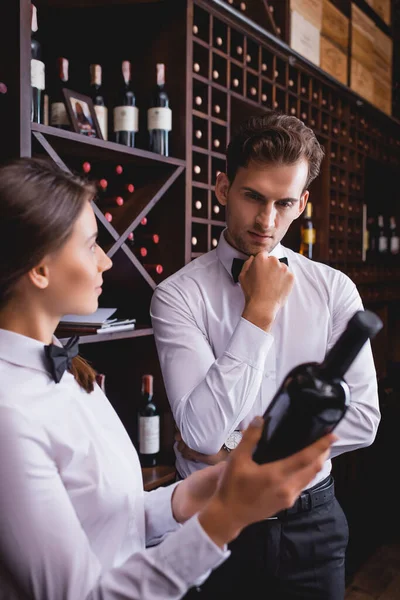 Селективный фокус задумчивого сомелье, стоящего рядом с коллегой с бутылкой вина — стоковое фото