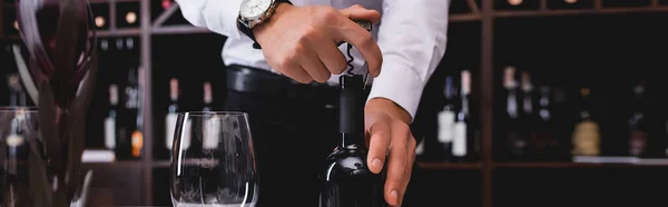 Panoramic shot of sommelier opening bottle of wine near glass of wine — Stock Photo