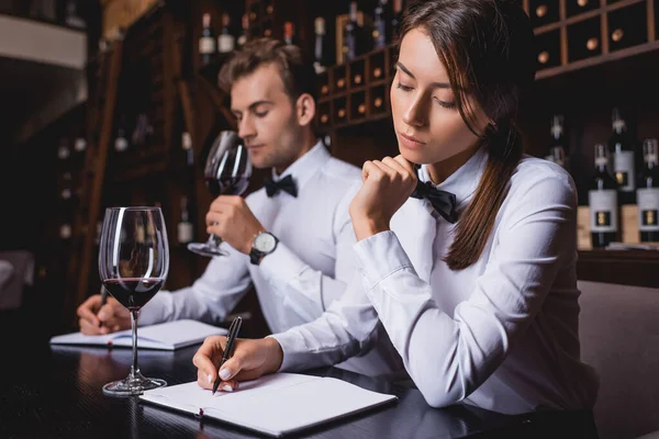 Enfoque selectivo de la escritura de sommelier concentrado en el cuaderno cerca de colega con vaso de vino - foto de stock