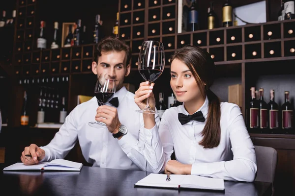 Selective focus of sommelier holding glass of wine near colleague and notebooks on table — Stock Photo
