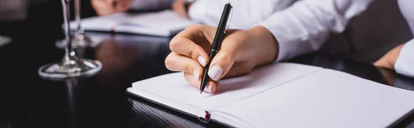 Panoramic shot of sommelier writing on notebook near glasses in restaurant — Stock Photo