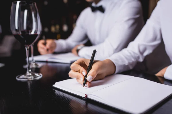 Vista cortada de sommelier escrevendo no caderno perto de colega e copos de vinho na mesa — Fotografia de Stock