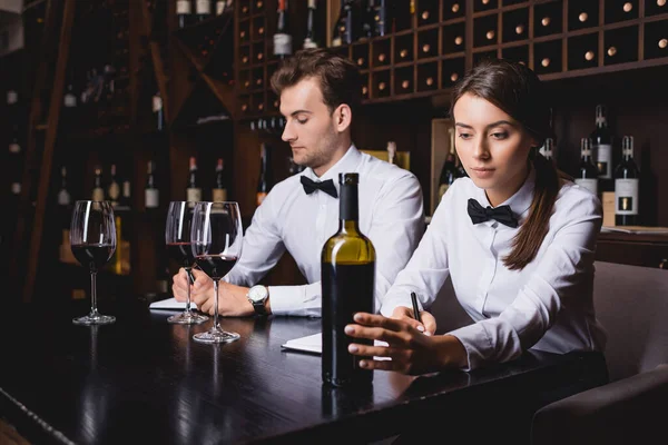 Selective focus of sommeliers noticing on notebooks near bottle and glasses of wine in restaurant — Stock Photo